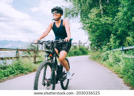 Portrait of a happy smiling woman dressed in cycling clothes, helmet and sunglasses riding a bicycle on the asphalt out-of-town bicycle path. Active sporty people concept image. Royalty-Free Stock Photo #2030336525
