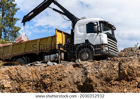 Side bottom pov big modern truck with mounted self loading crane arm boom unloading pallet of new bricks at countryside rural forest suburban construction site. Building materials delivery service Royalty-Free Stock Photo #2030352932