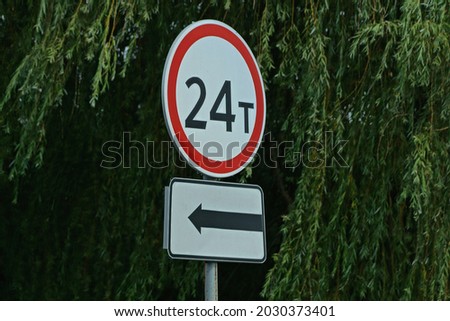 two road signs limiting the weight of 24 tons and an arrow on the street on a pole against a background of green vegetation