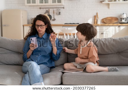 Focused on mobile phone woman working mother pay no attention on bored small kid. Young mom or nanny texting in smartphone sitting on sofa next to little son need parent love care and communication Royalty-Free Stock Photo #2030377265