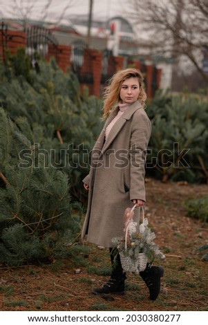 Elegant girl buys Christmas tree in a coniferous nursery. Girl near a Christmas tree on the street with a basket in hands, nature winter positive photo