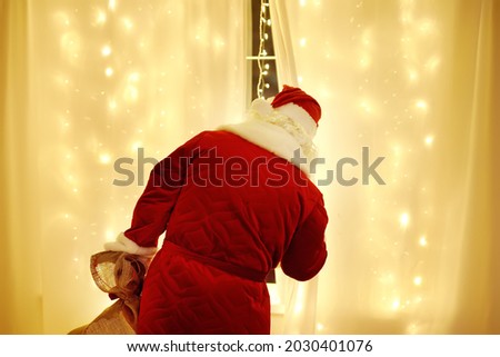 Santa Claus looks out window and waits for arrival of a sleigh pulled by deer. Animator or parent in Santa Claus costume is waiting for begin of holiday for children. Traditions around winter Holidays Royalty-Free Stock Photo #2030401076