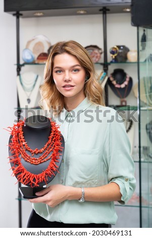 Successful young saleswoman demonstrating red coral necklace in jewelry shop.. Royalty-Free Stock Photo #2030469131