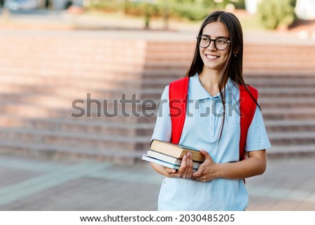 A smiling student girl with glasses, books and a red backpack. Portrait of a student on a city background. The concept of education