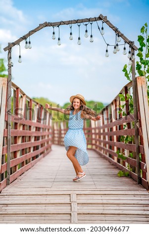 A happy little girl in a blue dress in a summer garden with a wooden bridge. Concepts of summer holidays and a fun childhood