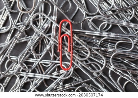 A red paper clip stands out against a textured background of silver paper clips. Concept