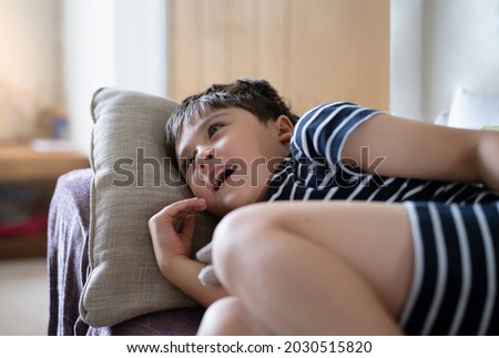 Happy boy lying on sofa looking up watching cartoon on TV. Cute Child resting in living room with bright light shining from window on sunny day summer. Kid relaxing at home on weekend 