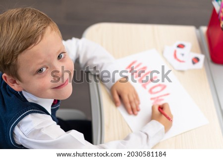 A first grader boy draws a postcard for the holiday of halloween with a red bright pencil on a white sheet of paper while sitting at a white table. Selective focus. Bokeh