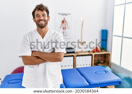 Young handsome physiotherapist man working at pain recovery clinic happy face smiling with crossed arms looking at the camera. positive person.  Royalty-Free Stock Photo #2030584364