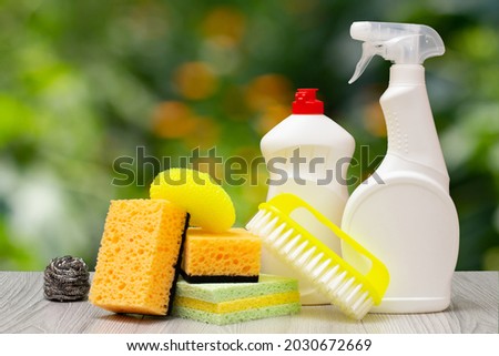 Plastic bottles of dishwashing liquid, glass and tile cleaner, a yellow brush and sponges on the blurred natural background. Washing and cleaning concept.