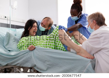 Pregnant woman in painful labor giving birth to child at maternity clinic. Obstetrician and black nurse assisting childbirth and helping young mother with delivery in hospital ward