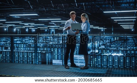 Data Center Female e-Business Enrepreneur and Male IT Specialist talk, Use Laptop. Two Information Technology Engineers on Bridge Overlooking Big Cloud Computing Server Farm. Royalty-Free Stock Photo #2030685665
