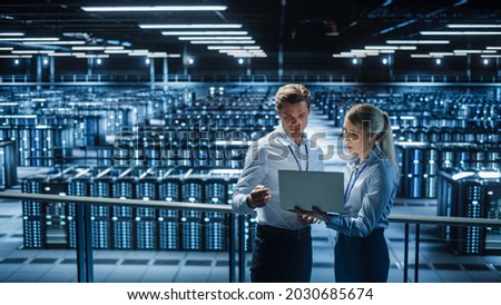 Data Center Female e-Business Enrepreneur and Male IT Specialist talk, Use Laptop. Two Information Technology Professionals on Bridge Overlooking Big Cloud Computing Server Farm. Royalty-Free Stock Photo #2030685674