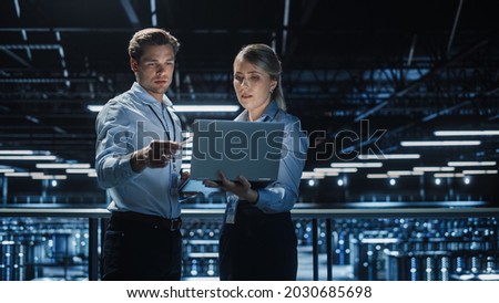 Data Center Female e-Business Enrepreneur and Male IT Specialist talk, Use Laptop. Two Information Technology Professionals on Bridge Overlooking Big Cloud Computing Server Farm. Royalty-Free Stock Photo #2030685698