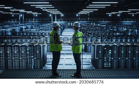 Data Center IT Specialist and System administrator Talk, Use Tablet Computer, Wearing Safety West. Server Cloud Farm Facility with Two Information Technology Engineers checking Cyber Security. Royalty-Free Stock Photo #2030685776