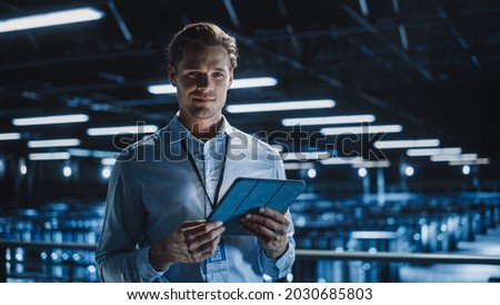Portrait of Handsome Smiling IT Specialist Using Tablet Computer in Data Center, Looking at Camera. Succesful Male e-Business Engineer Working in Big Server Farm Cloud Computing Facility. Royalty-Free Stock Photo #2030685803