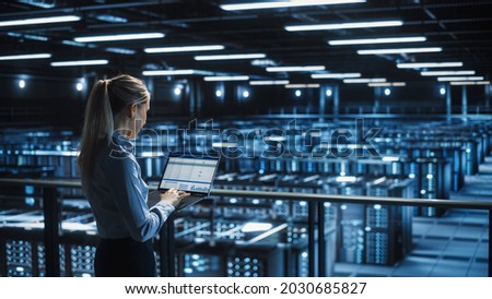 Data Center Female IT Specialist Uses Laptop Computer. Cloud Computing Server Farm with IT Engineer Monitoring Statistic, Maintenance Control. Information Technology of Fintech, e-Business. Royalty-Free Stock Photo #2030685827