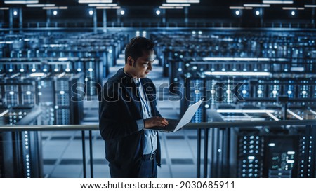 Portrait of IT Specialist Uses Laptop in Data Center. Server Farm Cloud Computing Facility with Male Maintenance Administrator Working. Cyber Security and Network Protection. Royalty-Free Stock Photo #2030685911