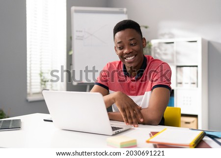 Trainee in office sits at desk in front of laptop monitor in background whiteboard, young dark-skinned man talks online with clients, co-workers, uses webcam, video call, smiling, cheerful Royalty-Free Stock Photo #2030691221