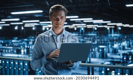 Handsome Smiling on Camera IT Specialist Using Laptop Computer in Data Center. Succesful Businessman and e-Business Entrepreneur Overlooking Server Farm Cloud Computing Facility. Royalty-Free Stock Photo #2030694491