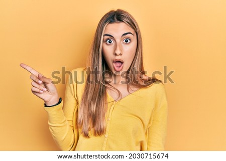 Beautiful hispanic woman wearing casual yellow sweater surprised pointing with finger to the side, open mouth amazed expression. 