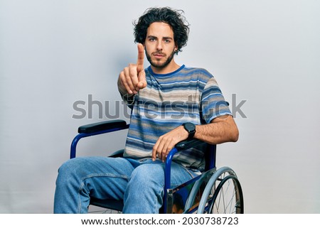 Handsome hispanic man sitting on wheelchair pointing with finger up and angry expression, showing no gesture 