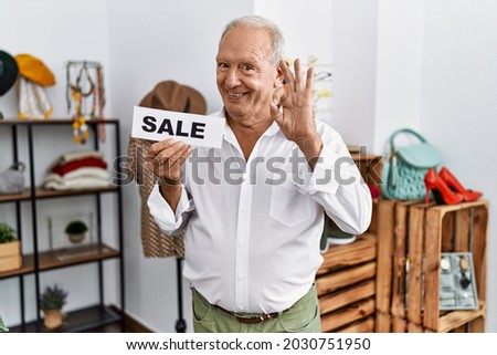 Senior man holding sale banner at retail shop doing ok sign with fingers, smiling friendly gesturing excellent symbol 