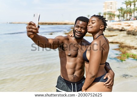 Young african american tourist couple wearing swimwear making selfie by the smartphone at the beach.