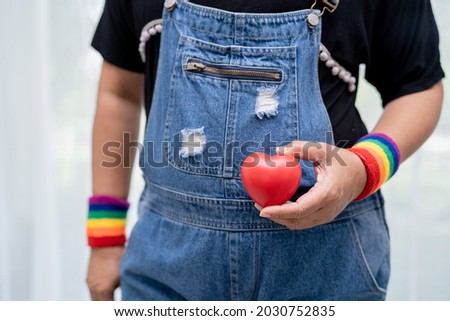 Asian lady wearing rainbow flag wristbands and hold red heart, symbol of LGBT pride month celebrate annual in June social of gay, lesbian, bisexual, transgender, human rights. Royalty-Free Stock Photo #2030752835