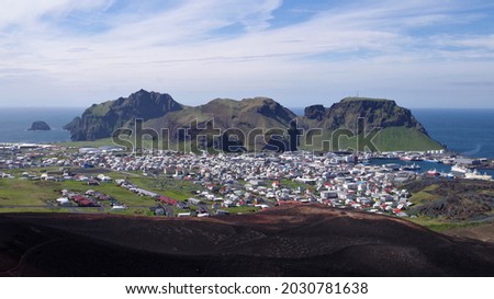 View over the town of Heimaey from neighbouring volcano, Eldfell. The sun shines on the buildings and the green and brown cliffs. Blue sea is visible in the background, and blue sky with white clouds.