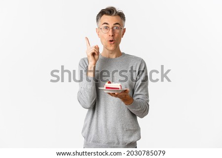 Image of happy middle-aged man, celebrating birthday, have great idea what to wish while blowing candle on b-day cake, standing over white background