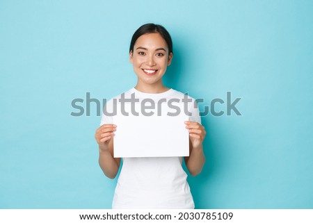 Waist-up portrait of smiling beautiful asian girl searching for someone, making announcement, holding blank piece of paper and looking at camera, standing light blue background