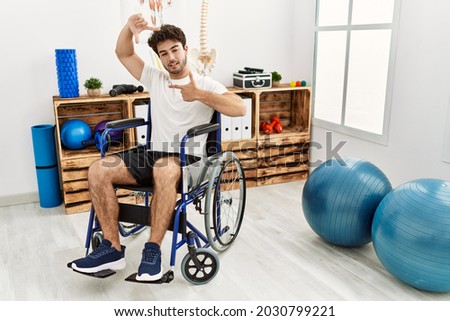 Hispanic man sitting on wheelchair at physiotherapy clinic smiling making frame with hands and fingers with happy face. creativity and photography concept. 