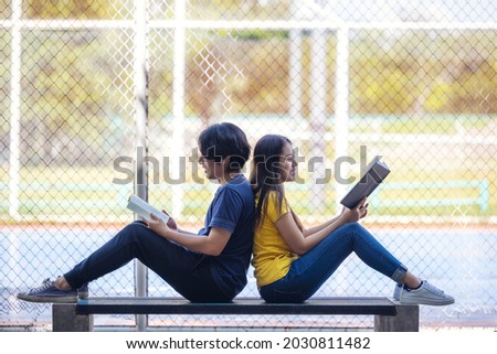 On a campus, a couple of students are studying together, and a teenager sits on a seat beside a sports court with a book.