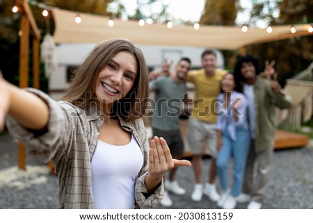 Lovely Caucasian woman taking selfie with her multiethnic friends next to motorhome on campsite, free space. Pretty young lady making mobile photo on autumn camping trip