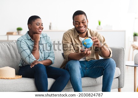 Cheerful Black Travelers Couple Posing With World Globe Choosing Destination For Next Vacation Sitting On Sofa At Home. Happy Tourists Planning Trip Together. Tourism And Traveling Concept