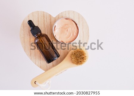 amber glass bottle with a pipette, a jar of cosmetics clay and a wooden brush for facial massage on a wooden heart-shaped stand, gray background, skin care concept