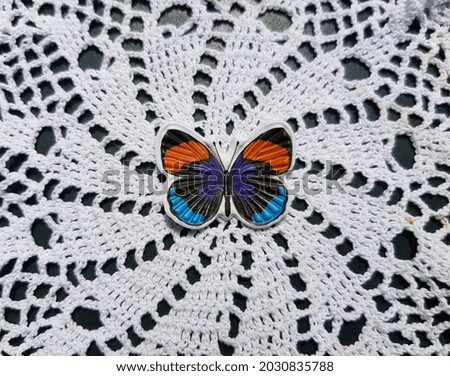 Small red, blue and black flat decorative butterfly on white knitted lace on a dark background (top view).