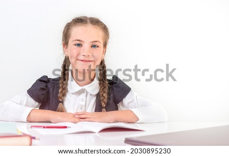 Schoolgirl sits behind a school desk and smiling on a white background