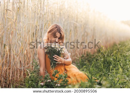 A young beautiful girl sits on a wheat field and holds a bouquet of daisies in her hands.