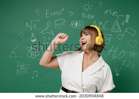 Happy excited teacher mature elderly senior lady woman 55 wears white shirt headphones singing isolated on green wall chalk blackboard background studio. Education in high school September 1 concept