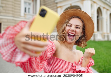 Young excited happy woman in pink dress hat do selfie shot on mobile cell phone post photo on social network do winner gesture walk in city center stand outdoor near building Urban lifestyle concept