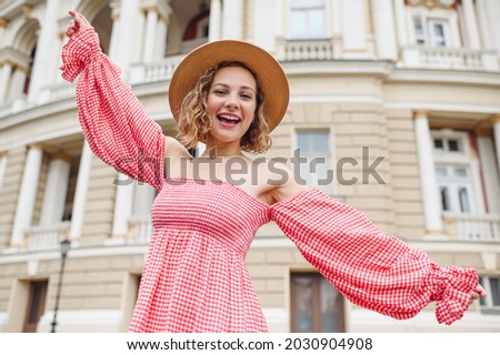 Young joyful smiling happy woman 20s wear pink dress hat dance with outstretched hands walk in city center standing outdoor near old building. People urban summer time lifestyle architecture concept