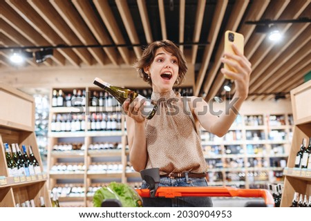 Young excited woman in casual clothes shopping at supermaket store with grocery cart hold white wine alcohol hold bottle do selfie shot on mobile phone inside hypermarket Purchasing gastronomy concept
