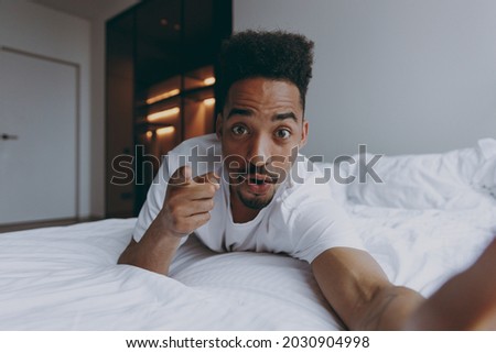 Close up young african american man in nightwear do selfie shot pov mobile phone point finger camera on you motivate do it lying in bed rest spend time in bedroom lounge home in own room house wake up