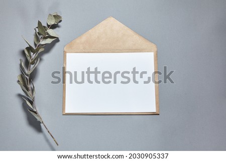 White paper sheet mockup on a brown craft envelope on gray background. White paper empty blank, branch of eucalyptus on gray table. Invitation card mockup. Flat lay, top view, copy space, mockup