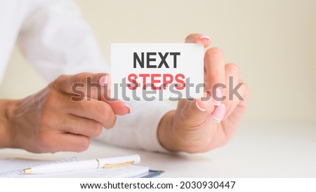 next steps inscription on white card paper sheet in hands of woman. black and red letters on white paper. business concept, grey backgrond