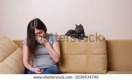 Young lady suffering from allergies sneezes into napkin sitting on sofa near gray cat at home
