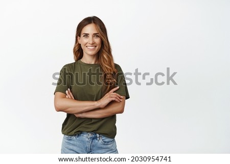 Portrait of confident adult woman in green t-shirt, standing with arms crossed on chest and self-assured motivated smile, looking at camera, standing against white background