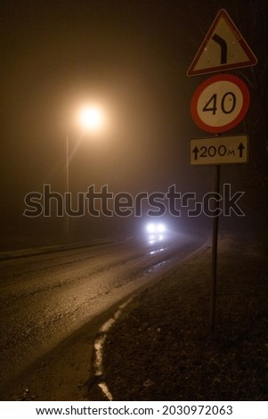 A road sign "dangerous turn" and "speed limit" against the background of car headlights and a street lamp on a foggy night